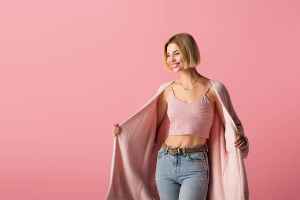 junge und zufriedene Frau in Strickjacke schaut vereinzelt auf rosa  - Foto, Bild