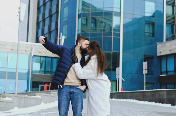 Love story of couple walking in city. Young business man in a blue jacket with a beard. And a cute woman in long coat. talking on the phone, making selfie. Bloggers. Family holiday, date - Foto, Imagem