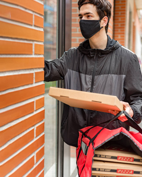 Yüzünde maske olan bir teslimatçı. Kapıcıyı arıyor, elinde eve teslim için kırmızı bir çanta tutuyor. - Fotoğraf, Görsel