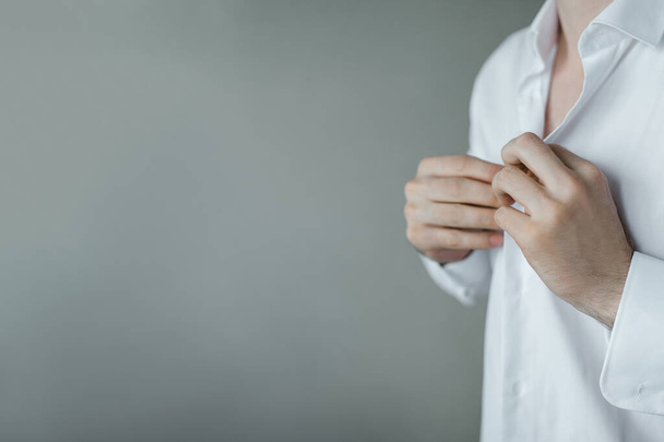 Ένας άντρας κουμπώνει τα κουμπιά ενός λευκού πουκάμισου στο φόντο του τοίχου - Φωτογραφία, εικόνα