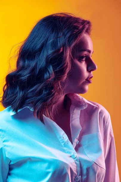 Profile view. Portrait of young beautiful girl with long curly hair posing isolated on orange background in neon light, filter. Concept of emotions, beauty, fashion - Photo, image