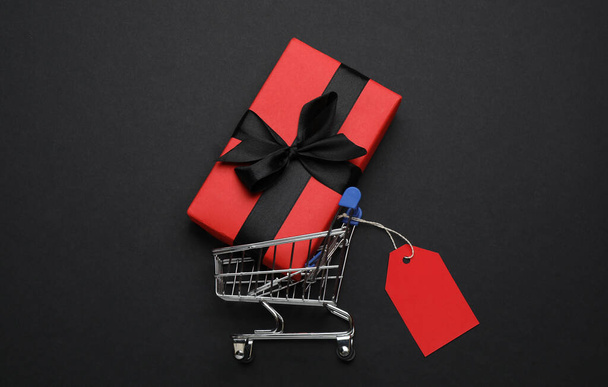 Shopping cart with price tag and gift box on black background, flat lay - Foto, Bild