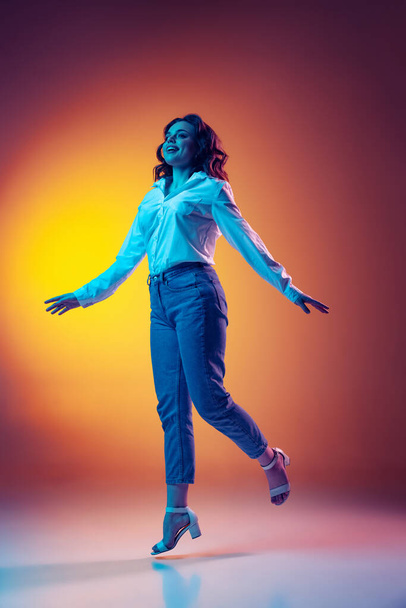 Full-length portrait of happy young beautiful girl with long curly hair jumping isolated on orange background in neon light, filter. Concept of emotions, beauty, fashion - Foto, Imagen