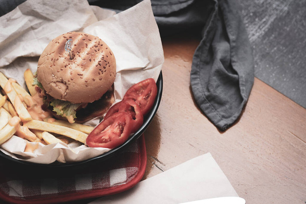 Kotitekoisia hampurilaisia tarjoillaan ranskalaisten perunoiden ja tomaattien kanssa puupöydällä. Pikaruokaa ja roskaruokaa. Modernilla äänensävyllä - Valokuva, kuva