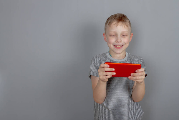 A laughing Caucasian boy in a gray T-shirt plays with a red smartphone and laughs on a gray background. Copy Space. - Foto, Imagen