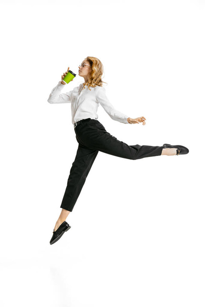 Dynamic portrait of young pretty girl wearing business style clothes jumping with coffee isolated on white background. Finance, ballet, art, business, beauty concept. - Foto, Imagen
