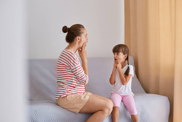 Horizontal shot of woman speech therapist wearing striped shirt and shorts sitting on sofa with little girl, proper articulation therapy for girl learning to speak correctly. - Фото, зображення
