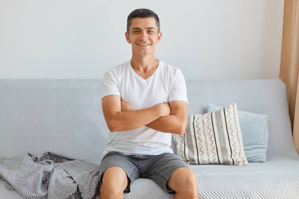 Indoor shot of smiling positive man wearing casual style white t shirt sitting on sofa, looking at camera, keeping hand folded, expressing positive emotions. - Foto, immagini