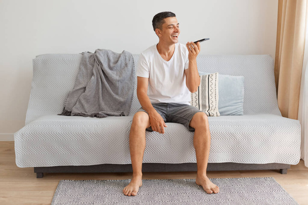 Indoor shot of attractive man wearing casual style white t shirt sitting on sofa and holding smart phone in hands and recording voice message, command for assistant. - Фото, изображение