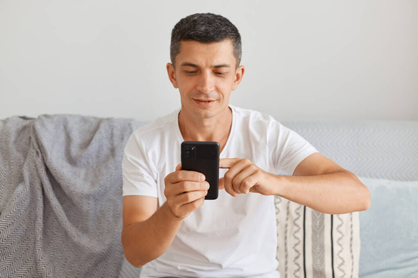 Image of smiling optimistic brunette man wearing casual style white t shirt sitting on cough and using cell phone, typing message, checking social networks, searching information in internet. - Photo, Image