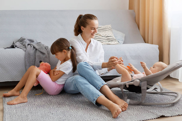 Portrait of smiling young adult woman spending time with her children at home, mother playing with infant baby, elder daughter feel jealous, sitting with upset expression. - Photo, image
