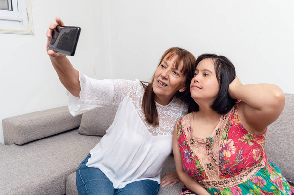 latinalaisamerikkalainen valkoihoinen aikuinen nainen, joka ottaa selfien tyttärensä kanssa, joka istuu kotona olohuoneen sohvalla, nuori nainen, jolla on alas-oireyhtymä äitinsä kanssa, joka ottaa valokuvia puhelimen elämäntapakonseptista - Valokuva, kuva