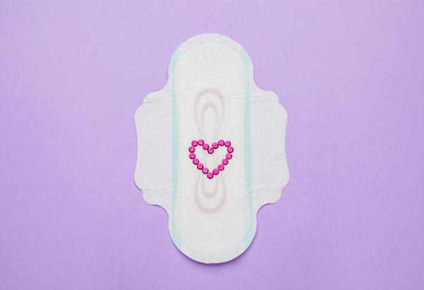Menstruatiepad met hartje gemaakt van roze pailletten en op violette achtergrond, bovenaanzicht - Foto, afbeelding
