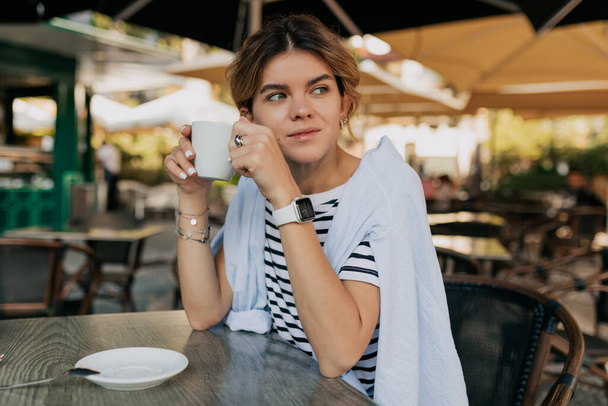 Adorável mulher adorável com cabelo recolhido vestindo t-shirt listrada e camisa azul sentado no café ao ar livre e beber café em dia ensolarado quente. Foto de alta qualidade - Foto, Imagem