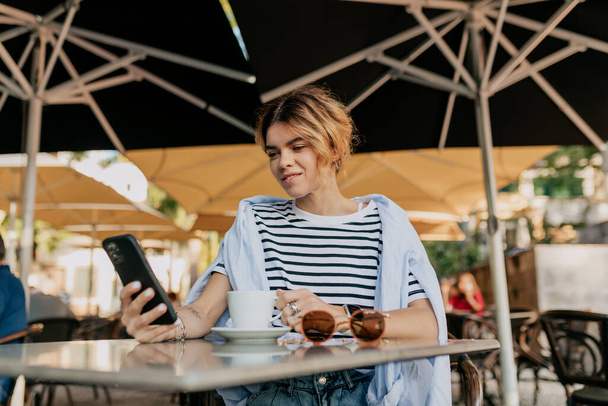 Entzückend hübsches Mädchen mit hellem Haar, gestreiftem T-Shirt und blauem Hemd sitzt in einer Cafeteria unter freiem Himmel und scrollt mit dem Smartphone. Glückliche Dame, die sich an sommerlich warmen Tagen gut amüsiert.  - Foto, Bild