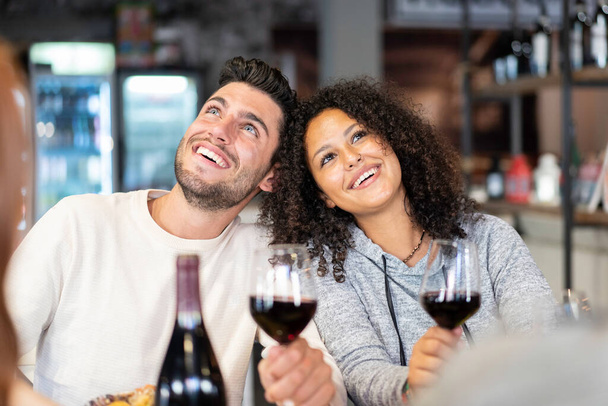 Happy multiracial couple of lover drinking red wine at fashion restaurant - Funny situation with both man and woman looking up - Relationship concept with boyfriend and girlfriend at first date - Φωτογραφία, εικόνα