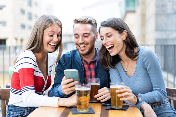 İnsanlar açık hava barında bira içerek eğleniyorlar - Genç arkadaşlarla birlikte vakit geçirip modern şehirdeki açık hava barında akıllı telefonlardan multimedya içeriğini izleyerek yaşam tarzı konsepti - Fotoğraf, Görsel