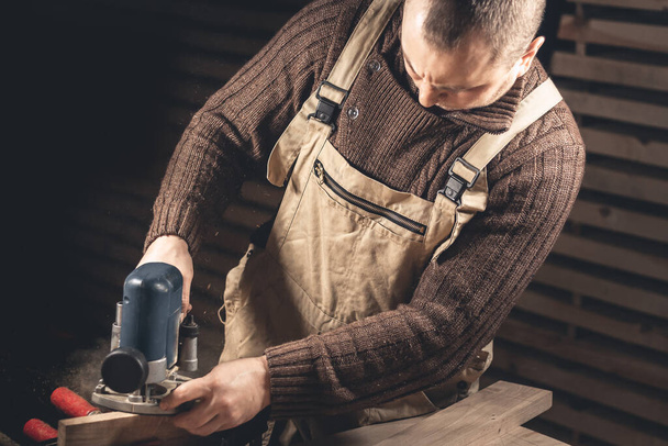 Ένας άνθρωπος φτιάχνει προϊόντα ξύλου με τη βοήθεια ειδικών εργαλείων. Πορτραίτο ενός νεαρού ξυλουργού στη δουλειά. Απασχόληση στην ξυλουργική βιομηχανία - Φωτογραφία, εικόνα