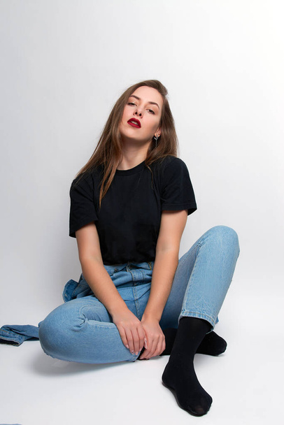 a girl with bright red lipstick, blue jeans and a black T-shirt sits on a white background - Photo, Image