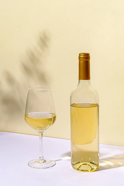 Botella sin etiquetar de vino blanco con media copa de vino vacía de pie sobre una mesa blanca contra una pared neutra con copyspace - Foto, Imagen