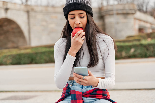 Zabawna głodna dziewczyna z apetytem gryzie czerwone jabłko siedzące w parku i patrzące na jej smartfon. koncepcja zdrowego życia - Zdjęcie, obraz
