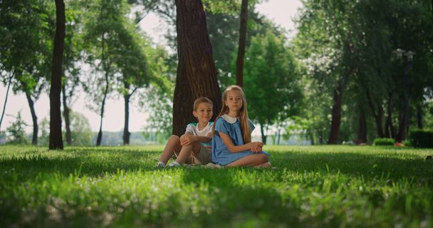 Usmívající se sourozenci si hrají na trávě pod stromem. Šťastné děti sedící na trávníku. - Fotografie, Obrázek