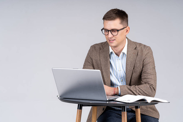 Handsome businessman sitting at the table and working at the laptop. Young male in suit is sitting and looking at the screen. Full length portrait - Foto, afbeelding