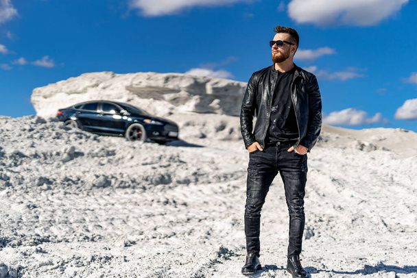Brutal man wearing suit is standing at the desert near his car. Man in front of the luxury auto. Horizontal photo - Foto, afbeelding