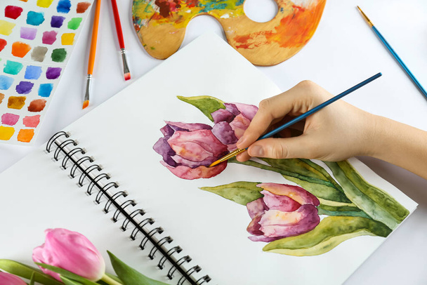 Woman painting tulips in sketchbook and supplies at white table, closeup - 写真・画像