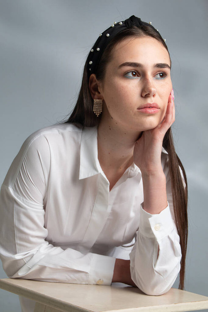 Waiting young woman with dark hair in white long sleeve blouse leaning on chair isolated on grey - Fotó, kép