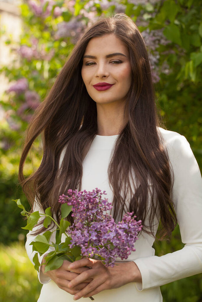 Linda jovem morena com longos cabelos ondulados vestindo vestido branco segurando buquê de flores da primavera - Foto, Imagem
