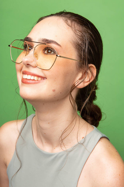 Portrait of smiling young woman with gathered dark hair in glasses isolated on green - Photo, image