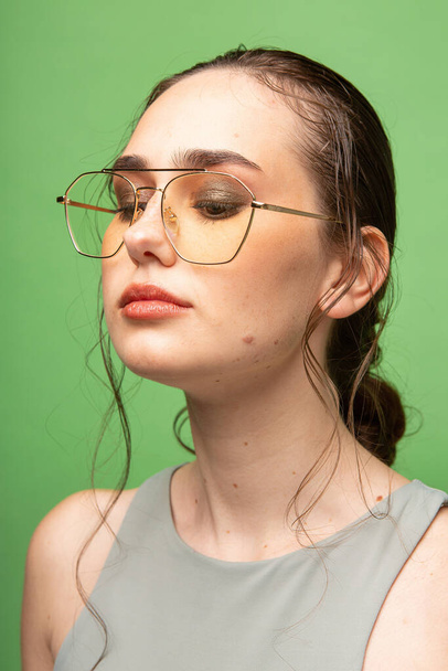 Portrait of looking down young woman with gathered dark hair in glasses isolated on green - Photo, image