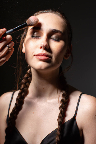 Portrait of making makeup smiling young european woman with two braids with closed eyes isolated on dark grey - Foto, Imagen