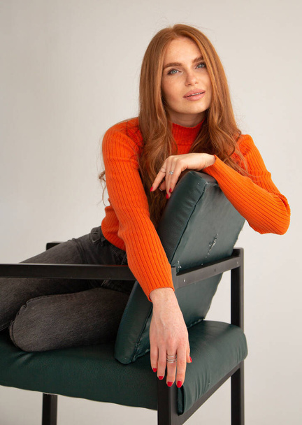 Woman with long red hair in orange jumper isolated on beige sitting on chair - 写真・画像
