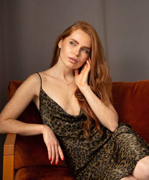 Woman with long red hair in animal print dress sitting on the couch in dark room - Photo, image