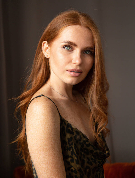 Portrait of woman with long red hair in animal print dress with gray eyes in dark room - Foto, afbeelding