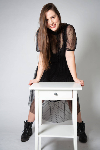 Young white european woman in black dress isolated on grey with white nightstand - 写真・画像