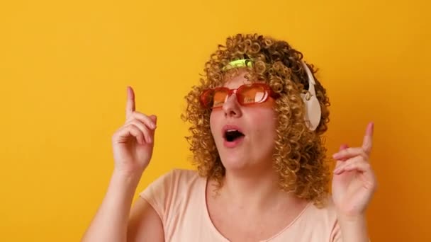 Funny cheerful wavy haired girl with glasses and a bandage listening soul dancing having fun isolated over yellow color background - Footage, Video