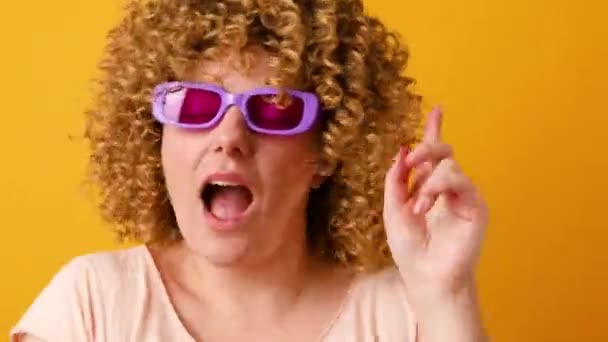 Šťastná optimistická mladá žena ve stylovém Veri pery barevné brýle zvedne paže a tance na oblíbenou hudbu, zapomněl na všechny problémy izolované přes žluté pozadí - Záběry, video