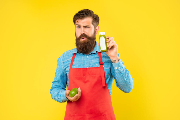 Winking man in apron holding limes and juice bottle yellow background, juice shopkeeper - 写真・画像