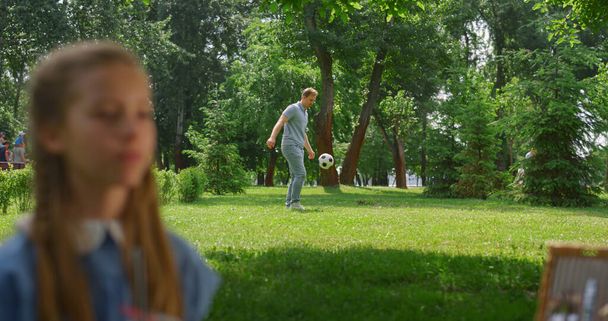 joven padre pasando pelota activo hijo detrás de descansando hija en parque. - Foto, imagen