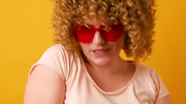 Happy young woman with curly fluffy hair in stylish love shape glasses raises arms and dances to favorite music isolated over yellow background - Footage, Video