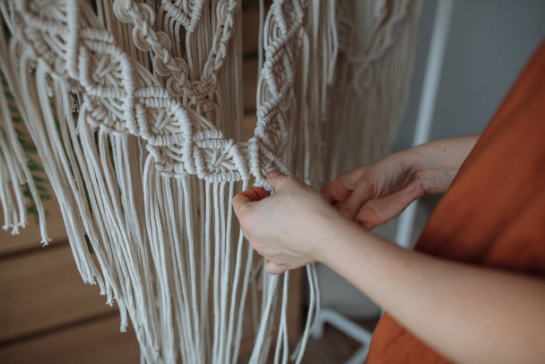 A young brunette girl in a brown shirt weaves a white panel at home using macrame technique - Фото, изображение