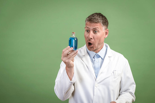 Scientist in white coat holding small flask with blue liquid and is standing in front of green background - Fotoğraf, Görsel