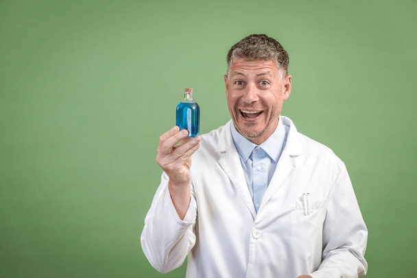 Scientist in white coat holding small flask with blue liquid and is standing in front of green background - Fotoğraf, Görsel