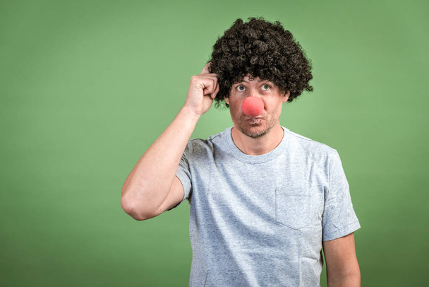 Man with black wig and red clown nose posing in front of green background - Фото, зображення
