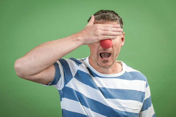 Man with red clown nose and gray hair with blue and white striped shirt stands in front of green background in the studio - Photo, Image