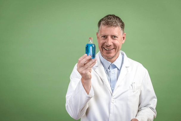Scientist in white coat holding small flask with blue liquid and is standing in front of green background - Фото, зображення