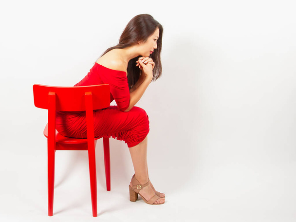 Mujer asiática pensativa con el pelo largo, morena, en un vestido elegante rojo con escote, se sienta en una silla roja y mira hacia abajo. - Foto, imagen